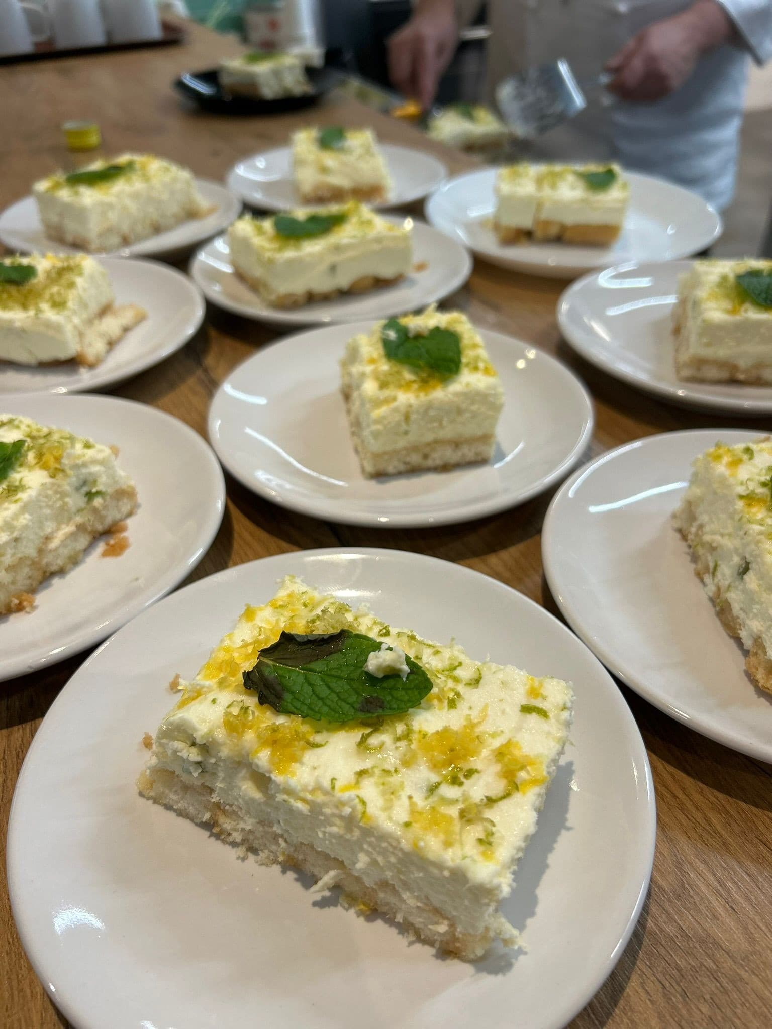 Mexicaanse avond: nagerecht per portie op witte bordjes, met roomlaag, fijngeraspte citrus en munt; kok in witte koksjas serveert op de achtergrond.